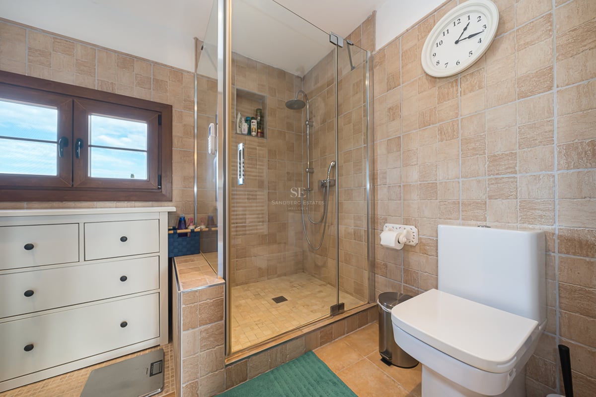 Salle de bain avec carrelage beige texturé, douche vitrée, commode blanche et fenêtre en bois.