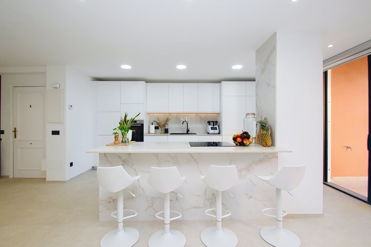 Modernt vitt kök med en stor köksö i marmor, fyra vita barstolar och inbyggda vitvaror.