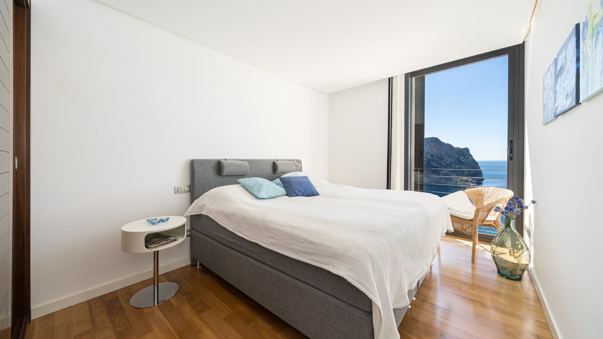 Contemporary bedroom with a gray upholstered bed, wooden floors, and a floor-to-ceiling window overlooking the sea.