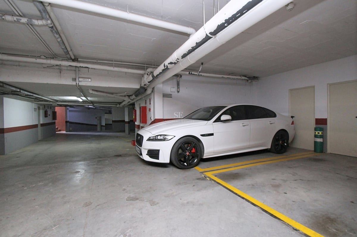 En vit bil parkerad i ett ljust underjordiskt garage med betonggolv och gula parkeringslinjer.