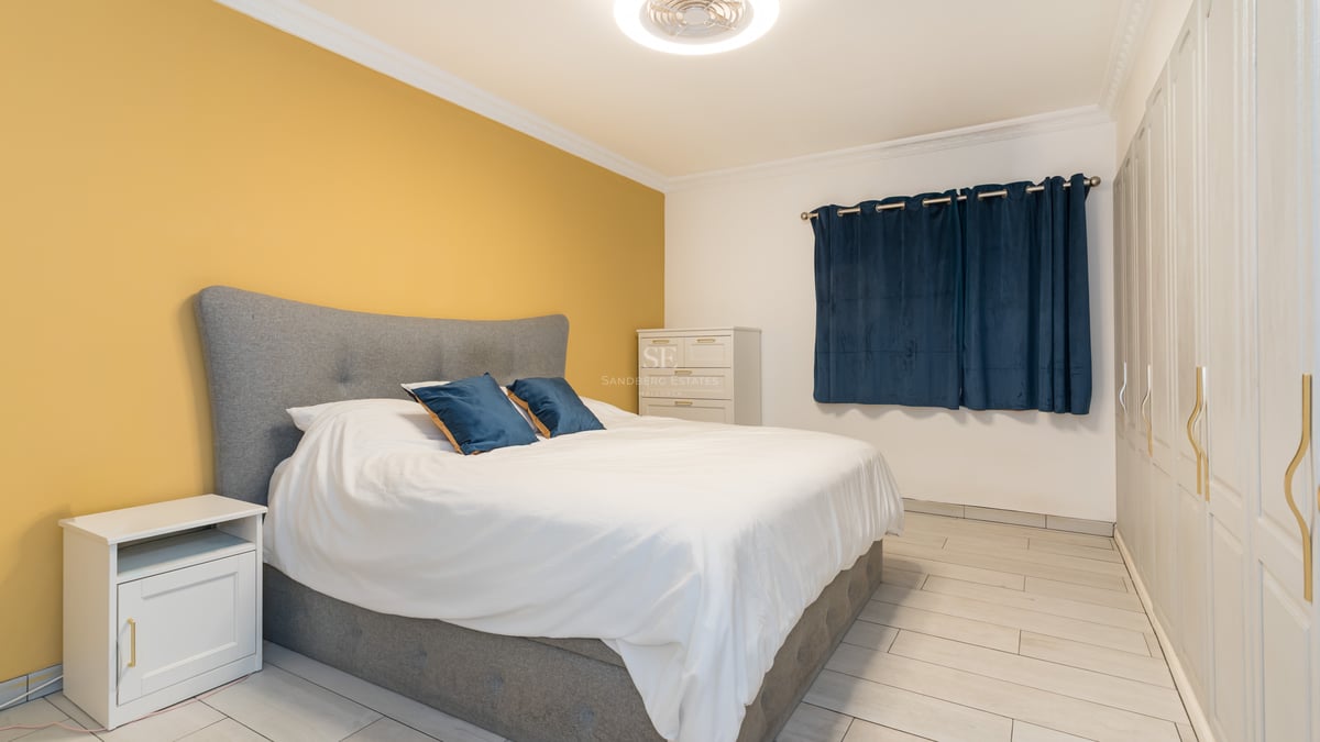 Une chambre lumineuse avec lit gris, mur jaune, placards blancs et rideaux bleu foncé.