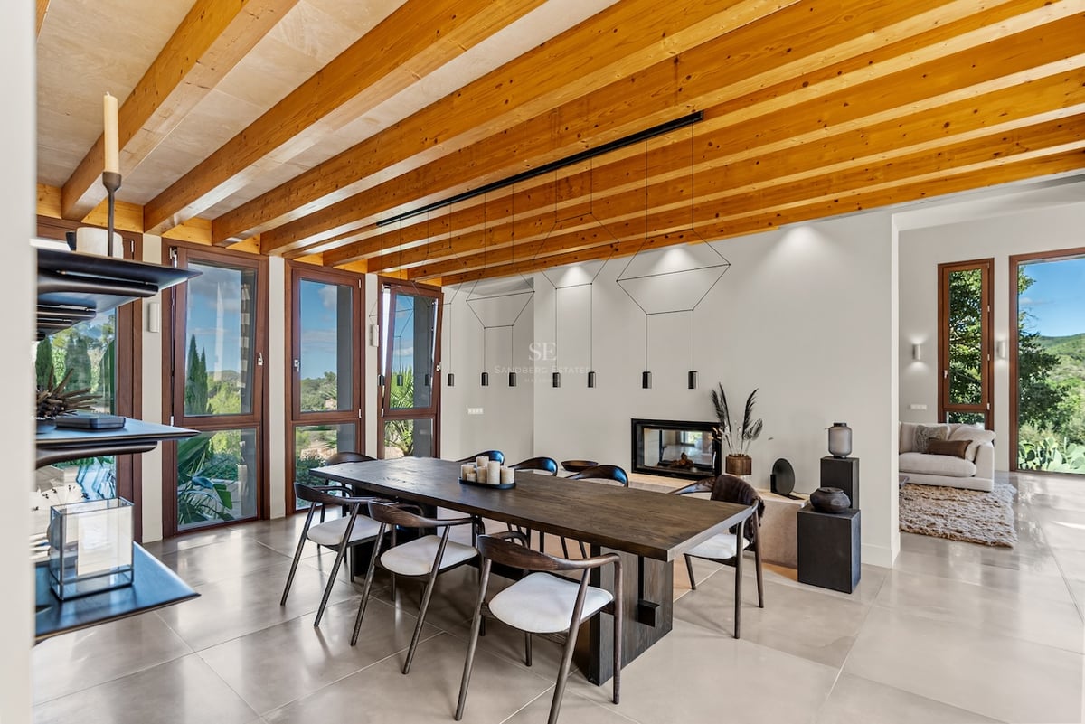 Grande table à manger en bois sous un plafond à poutres avec des baies vitrées donnant sur la nature.