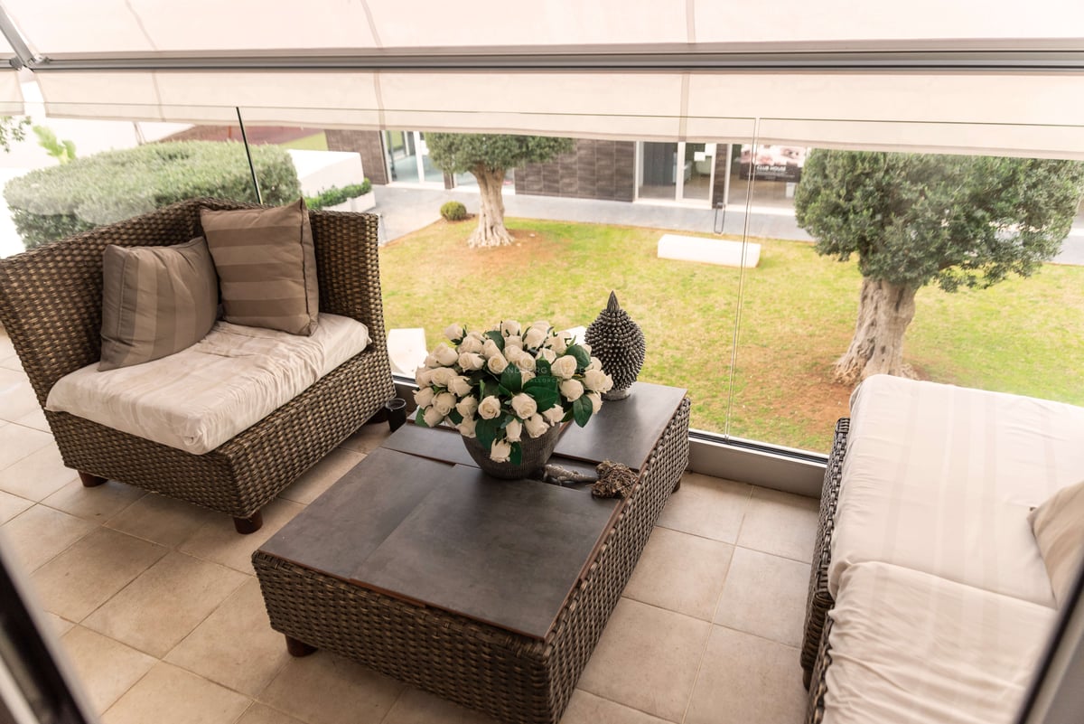 Überdachte Terrasse mit Rattan-Loungemöbeln, Glasgeländer und Blick auf einen Garten mit Olivenbäumen.