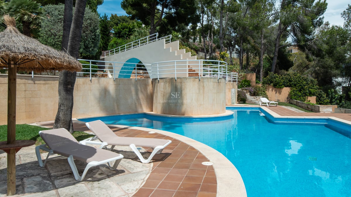 Large blue tiled swimming pool with sun loungers, a straw umbrella, and a decorative white bridge structure in a garden.