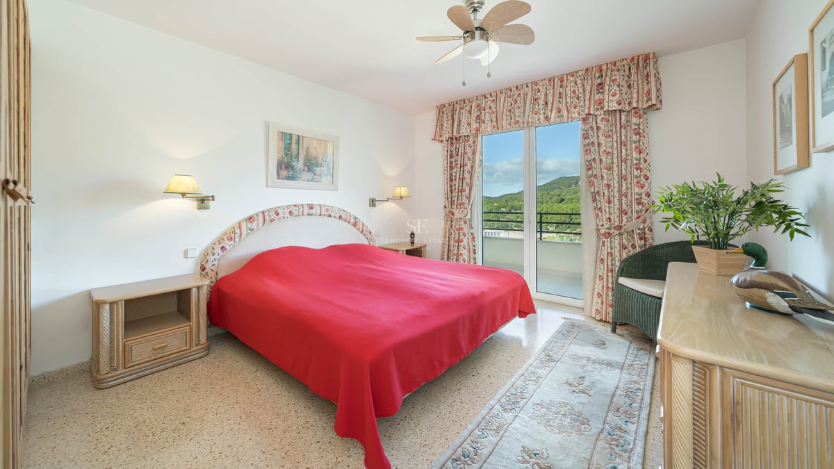 Bright bedroom featuring a red bedspread, floral curtains, and glass doors opening to a balcony with green hill views.