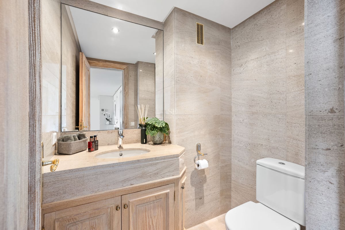 Salle de bains moderne avec carrelage en pierre beige, grand miroir et meuble sous vasque en bois.