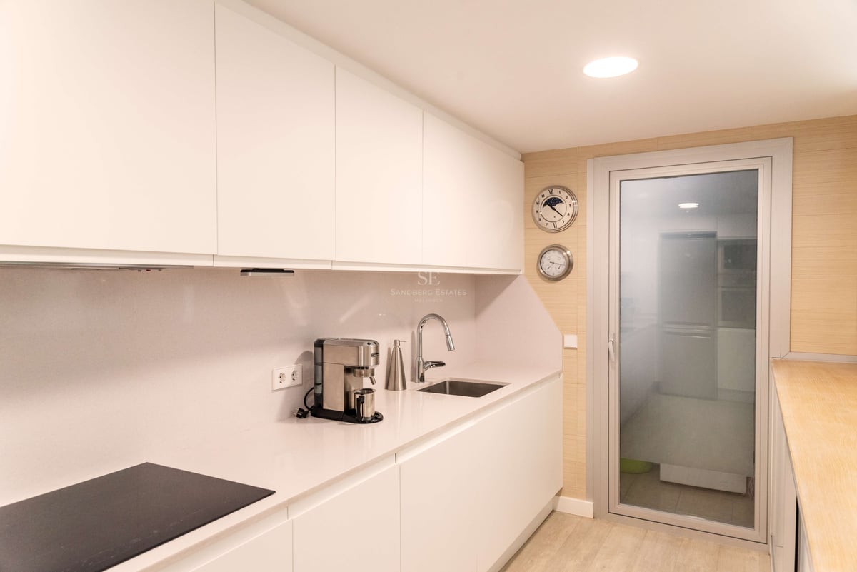 Moderne weiße Küche mit grifflosen Schränken, Quarz-Arbeitsplatte und Glastür zum Außenbereich.