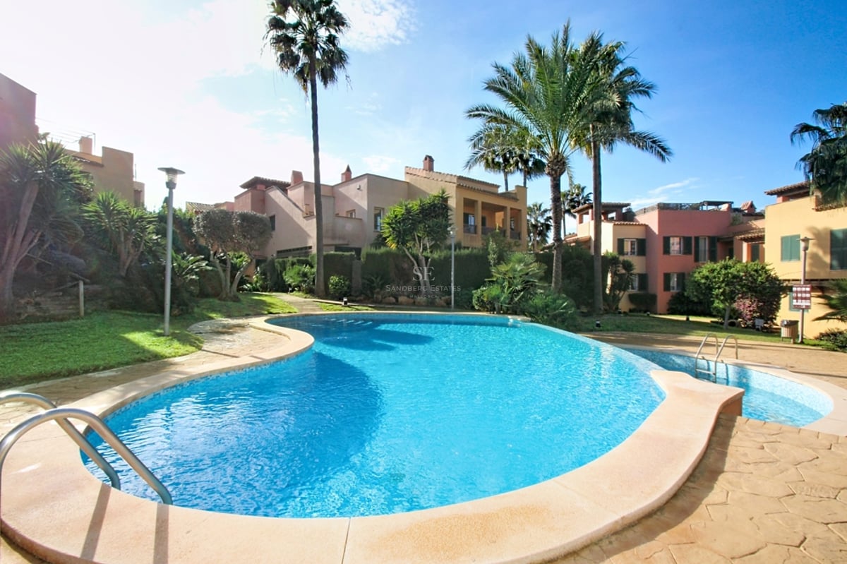 Oregelbundet formad pool omgiven av tropiska trädgårdar och byggnader i medelhavsstil i dagsljus.