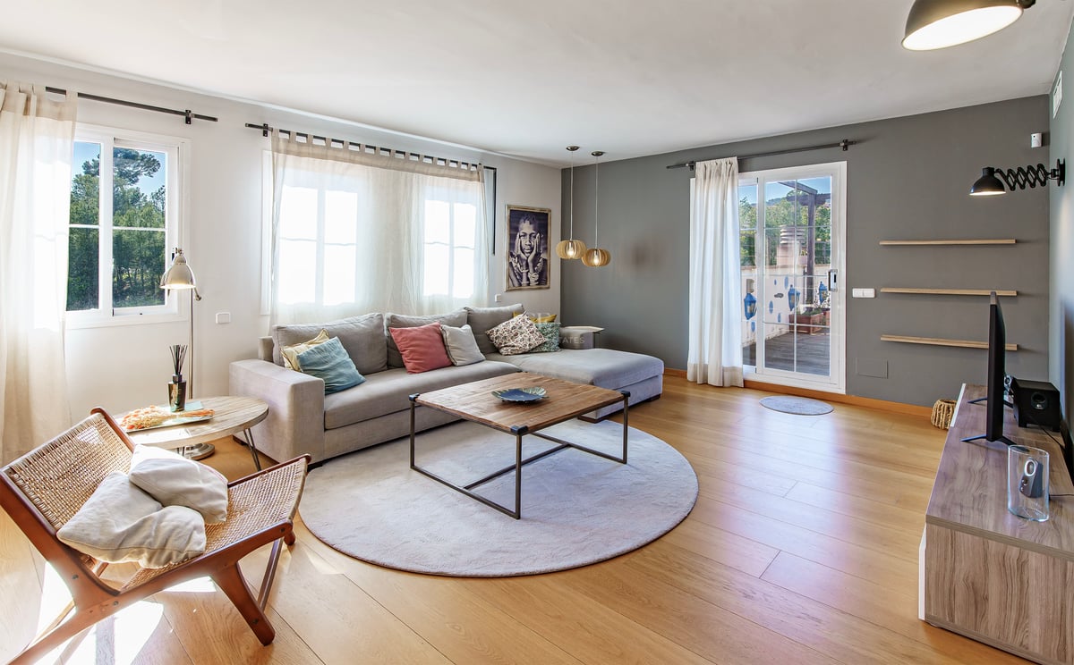 Ljust vardagsrum med grå hörnsoffa, ekparkett och glasdörr som leder till en solig terrass.