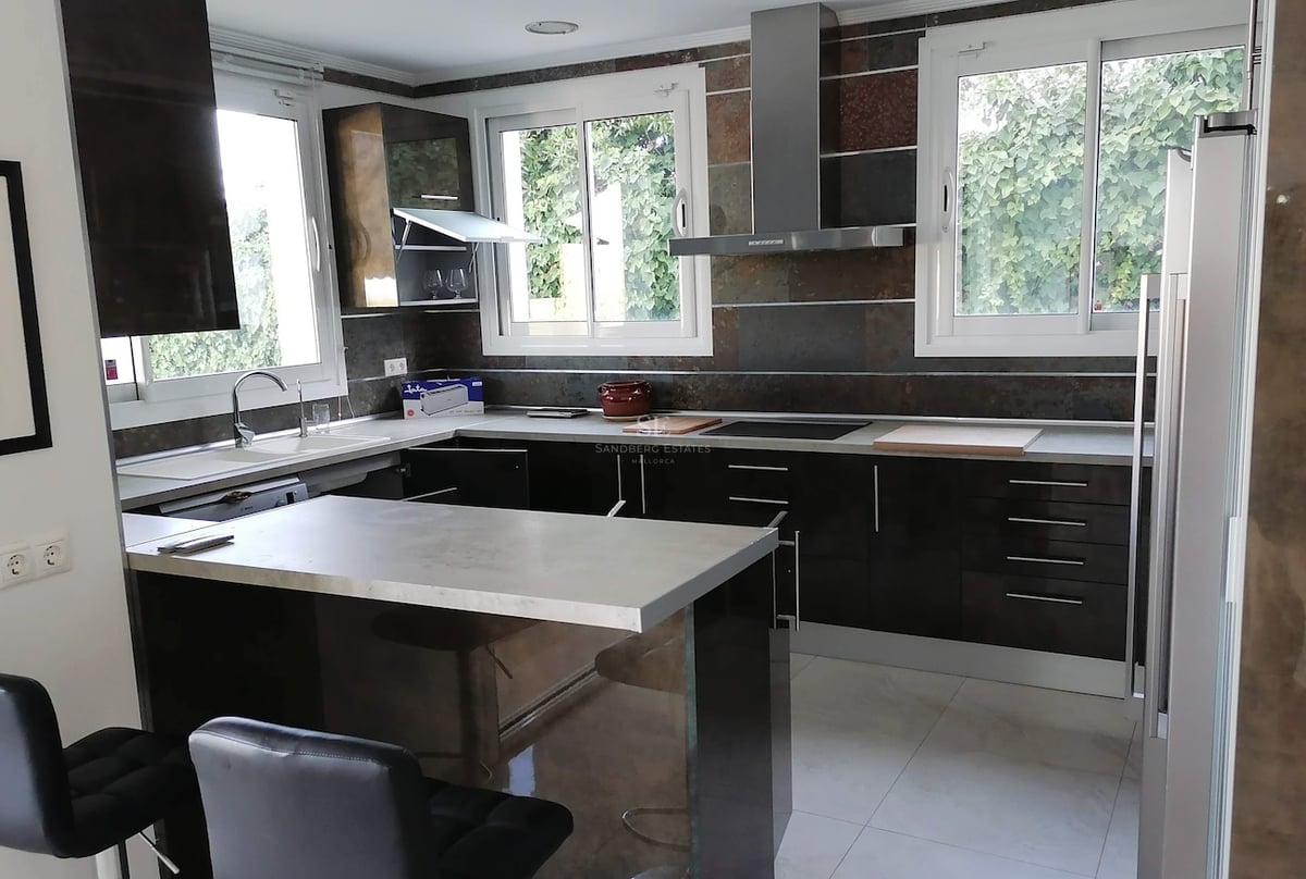 Cuisine moderne en L avec placards sombres brillants, plan de travail gris et fenêtres donnant sur le jardin.