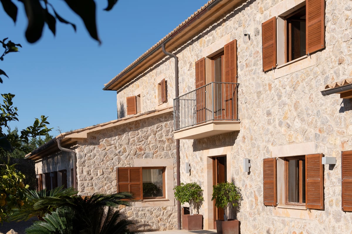 Exteriör av en tvåvånings stenvilla med fönsterluckor i trä och en balkong under en klarblå himmel.