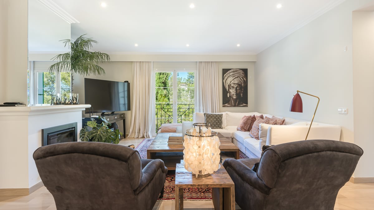 Bright living room with white L-shaped sofa, dark armchairs, wooden coffee table, and balcony overlooking greenery.