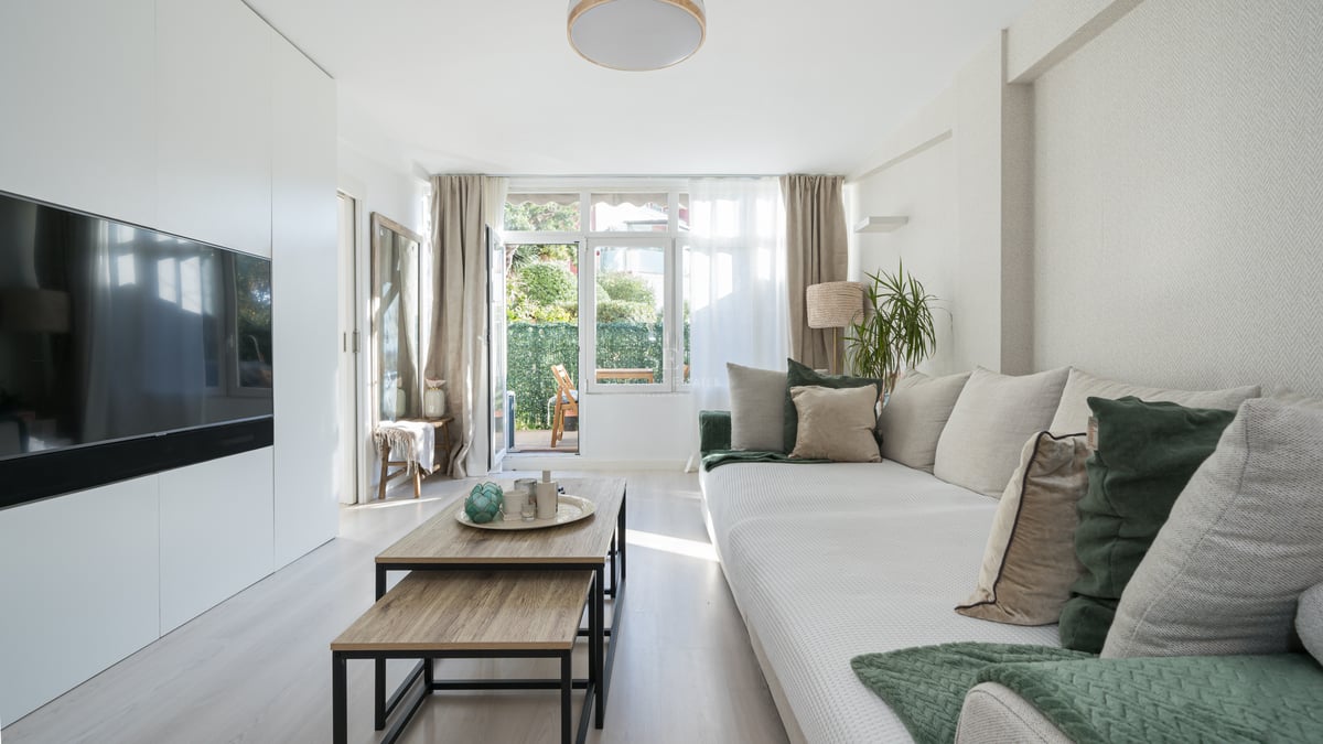 Bright living room featuring a large beige sofa, wooden coffee tables, and glass doors opening to a green terrace.