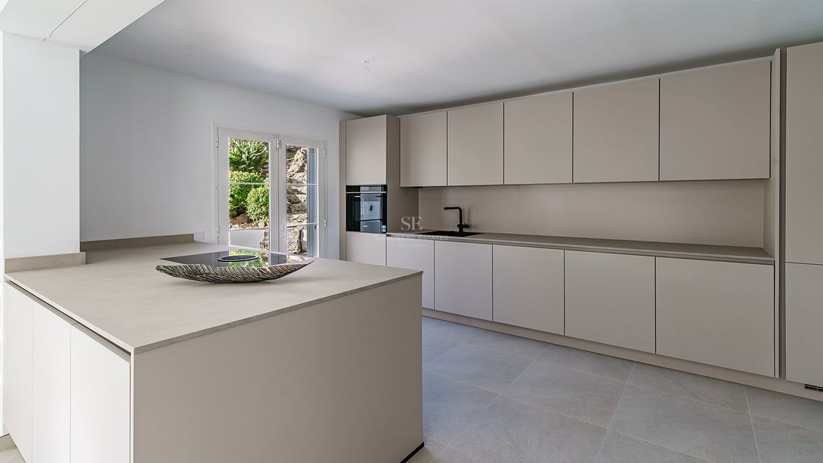 Samtida kök med sandfärgade handtagslösa skåp, central köksö och utgång till trädgård.