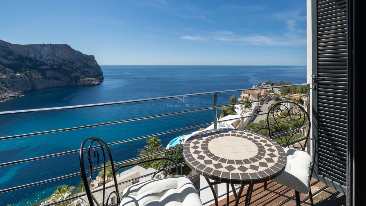 A small terrace with a mosaic table and iron chairs overlooking a deep blue Mediterranean bay and rugged cliffs.