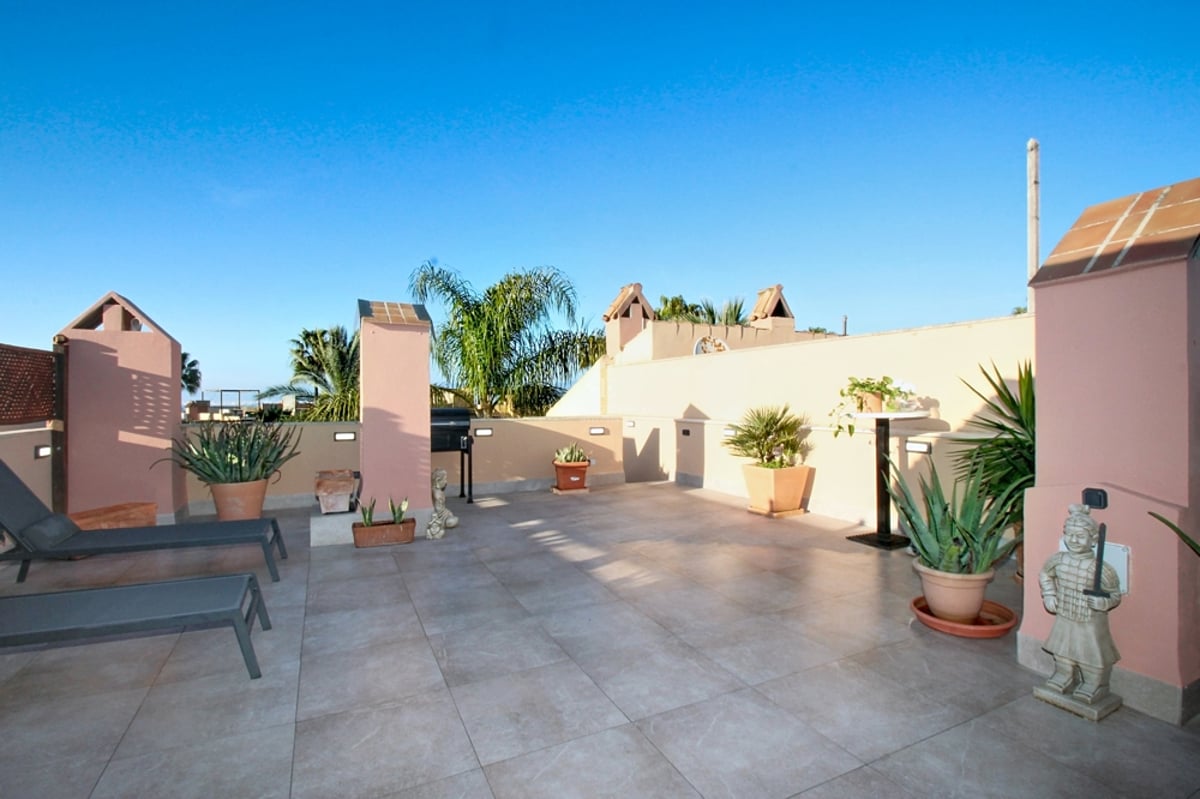 Stor takterrass med beige klinker, rosa putsade väggar, solstolar, krukväxter och klarblå himmel.