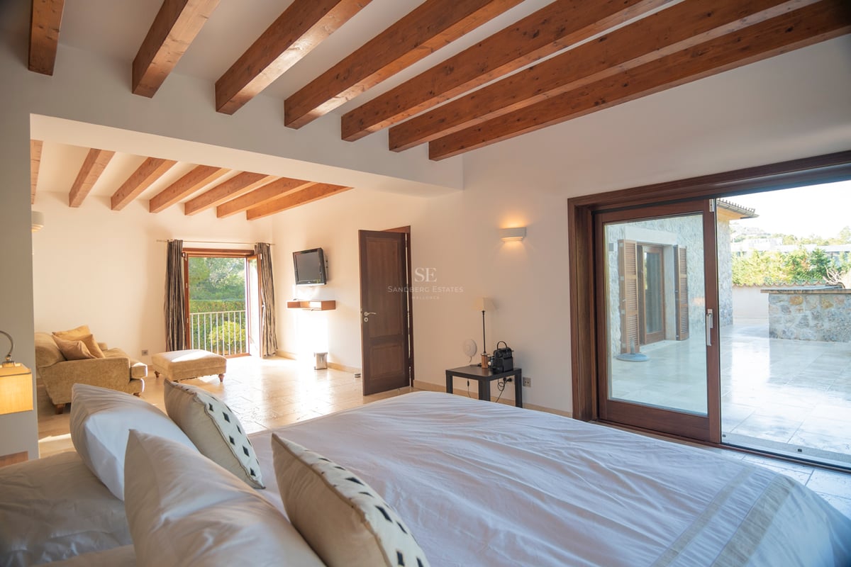 Rymligt sovrum med högt i tak, takbjälkar i trä, stengolv och skjutdörrar i glas mot terrassen.