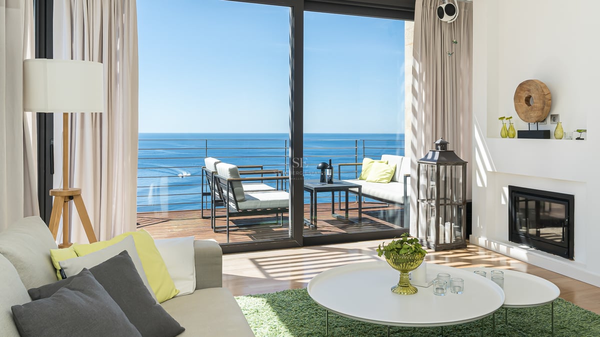 Contemporary living room with white decor, fireplace, and floor-to-ceiling glass doors opening to a sea-facing terrace.