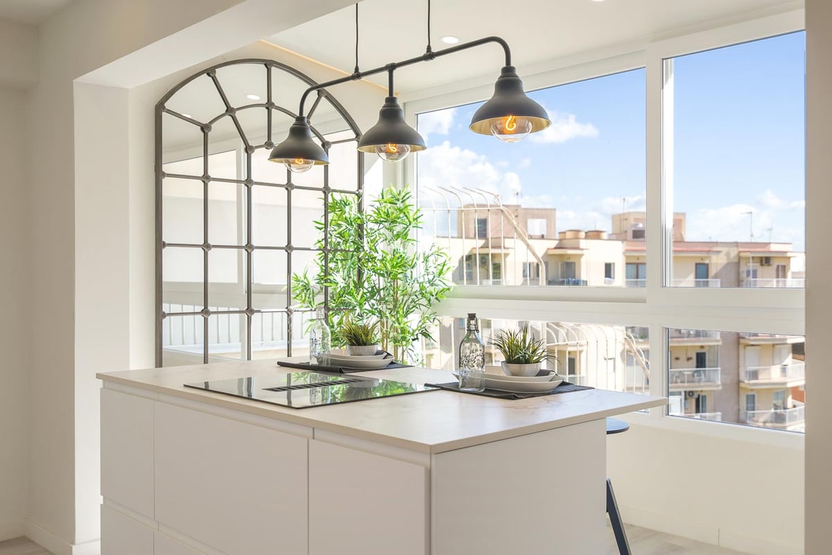 Weiße Kücheninsel mit Induktionskochfeld, Industrielampen und großen Fenstern mit Blick auf Stadtgebäude.