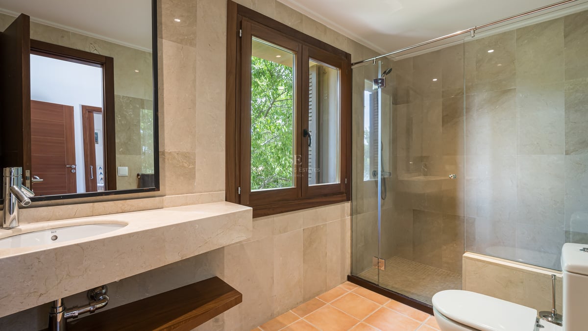 Salle de bain avec douche à l'italienne vitrée, carrelage en pierre beige et fenêtre en bois sur le jardin.