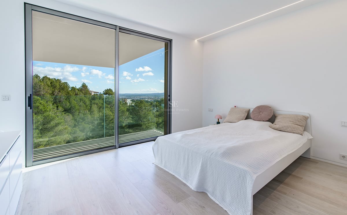 Chambre minimaliste lumineuse avec de grandes baies vitrées ouvrant sur un balcon avec vue sur les collines verdoyantes.