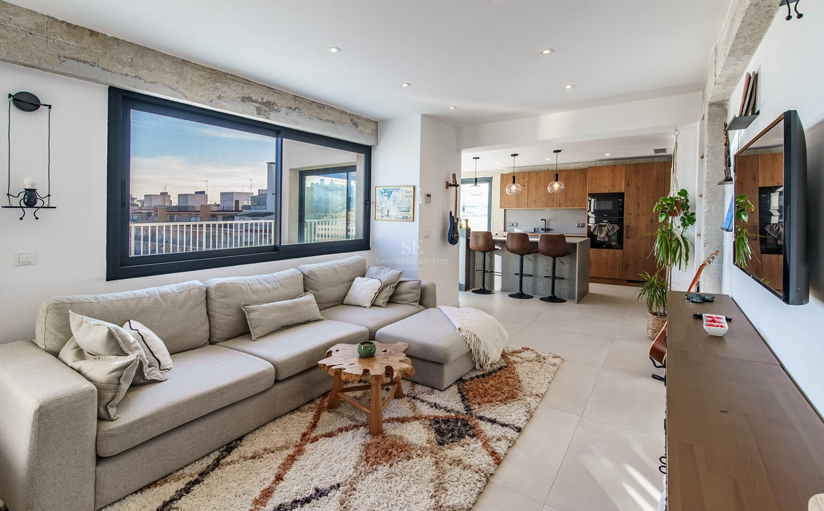 Salon moderne et lumineux avec un grand canapé d'angle gris, un tapis texturé et une cuisine ouverte avec vue sur la ville.