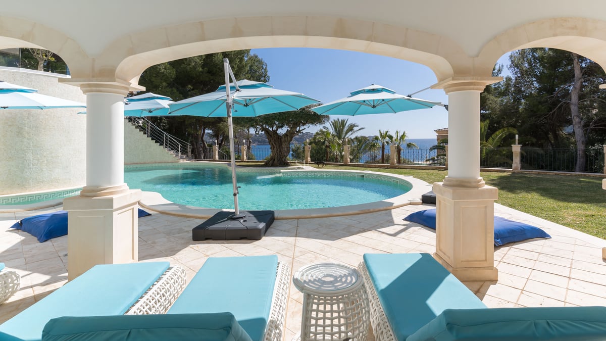 Ein geschwungener türkisfarbener Swimmingpool, umgeben von Natursteinfliesen und türkisfarbenen Liegen unter einem Torbogen.