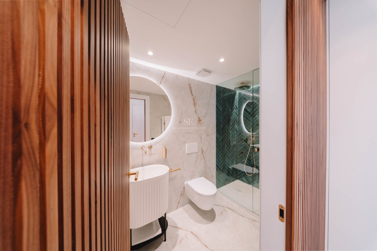 Salle de bain moderne avec marbre, miroir rond rétroéclairé, robinetterie dorée et douche en chevrons verts.