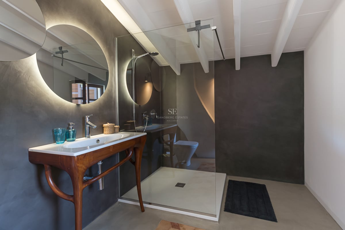 Salle de bain moderne avec murs en microciment gris foncé, miroir circulaire rétroéclairé et meuble en bois sculpté.