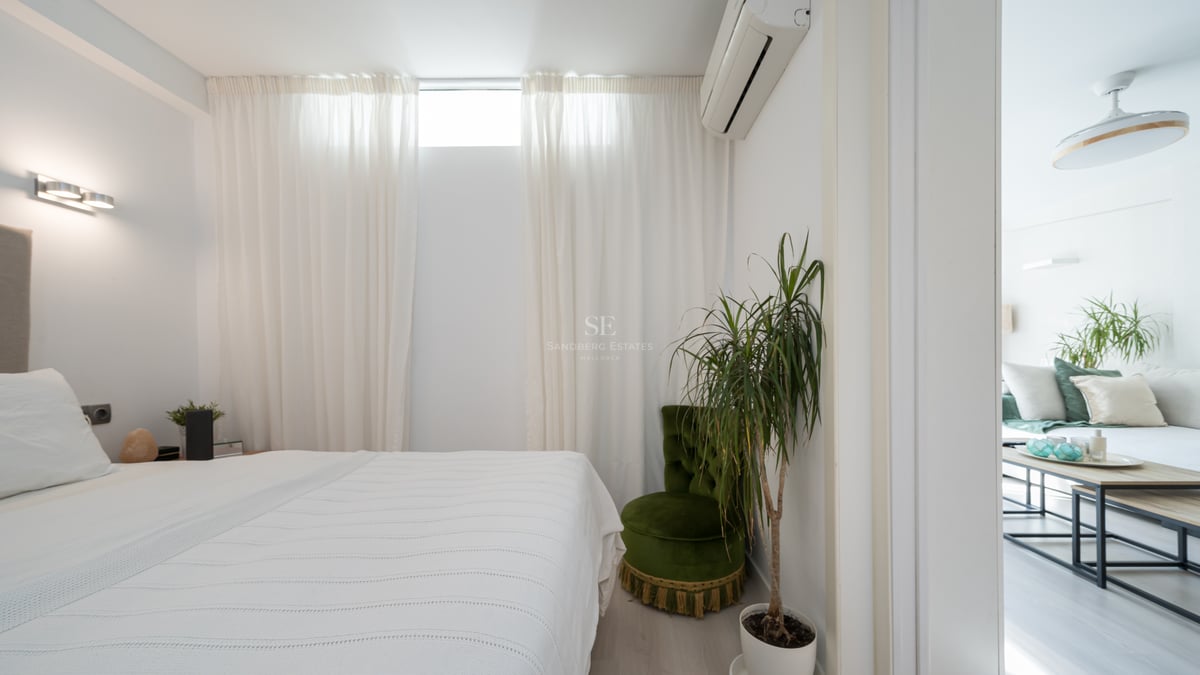 Clean white bedroom featuring a bed, green velvet chair, indoor plant, and a doorway looking into the living room.