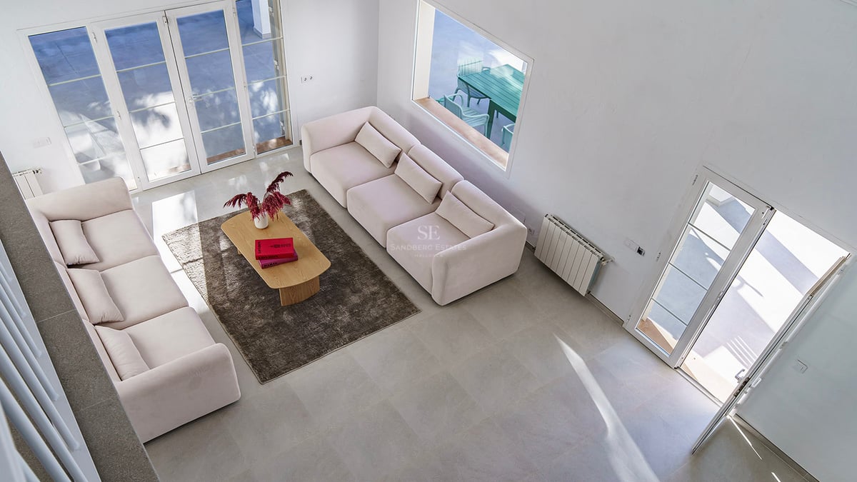 Upphöjd vy av ett ljust vardagsrum med cremefärgade soffor, soffbord i trä och stora glasdörrar mot en terrass.