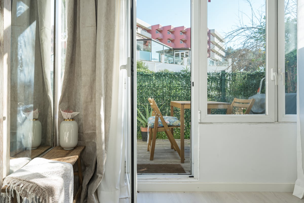 View through a glass door to a wooden terrace with outdoor furniture and a privacy hedge.
