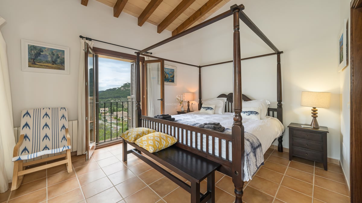 Chambre principale avec lit à baldaquin, poutres en bois, sol en terre cuite et balcon donnant sur les collines.