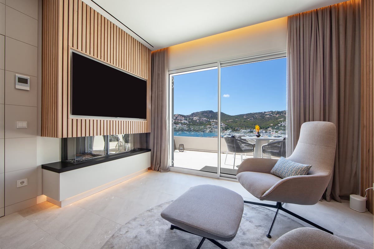 Salon contemporain avec fauteuil, cheminée, télévision sur mur en bois et vue panoramique sur le port depuis le balcon.