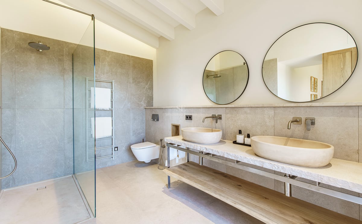 Luxuriöses Badezimmer mit zwei Steinwaschbecken auf Marmorplatte, Rundspiegeln und begehbarer Glasdusche.