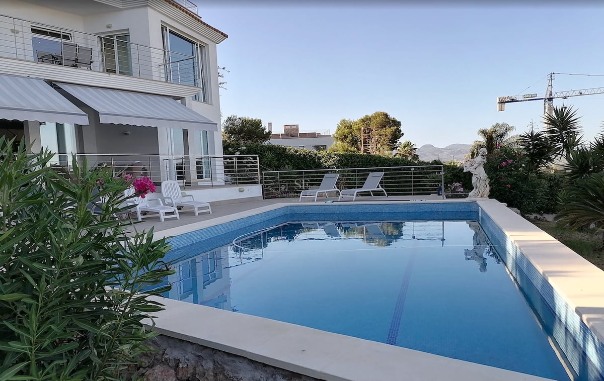 Une piscine rectangulaire en mosaïque bleue devant une villa blanche avec des chaises longues et des plantes.