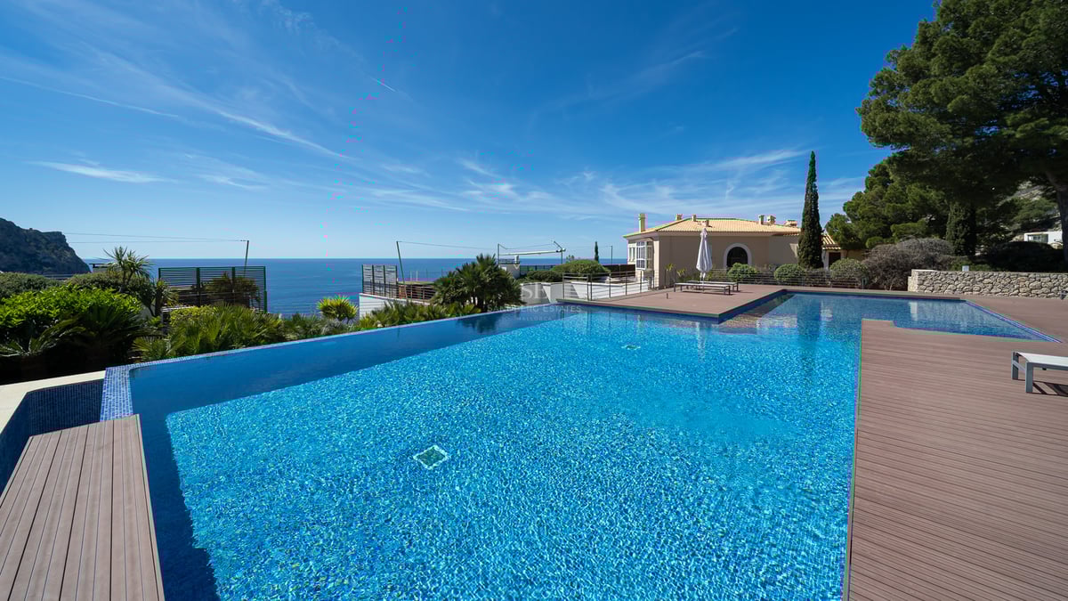 Large infinity swimming pool with blue tiles overlooking the sea and a stone terrace under a clear sky.