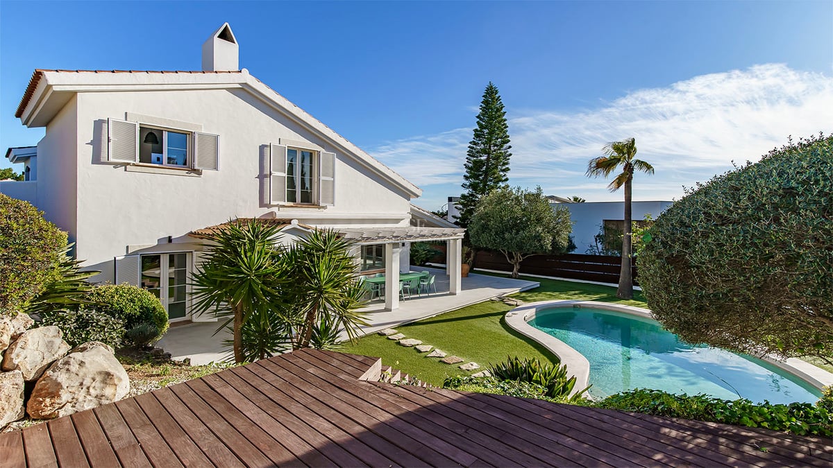 Vit tvåvåningsvilla med en njurformad pool, trädäck och välskött trädgård under klarblå himmel.