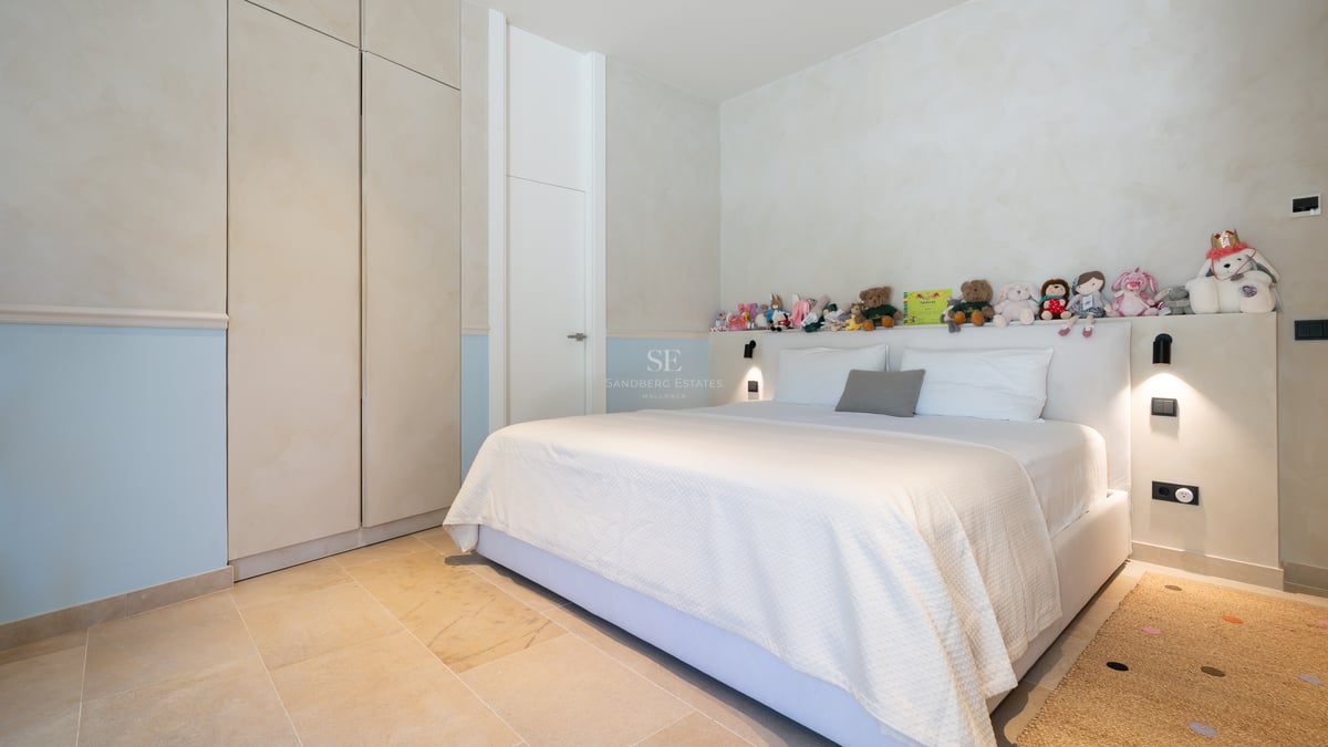 Chambre lumineuse avec un grand lit, linge de lit blanc, sol en pierre naturelle et placards intégrés crème.