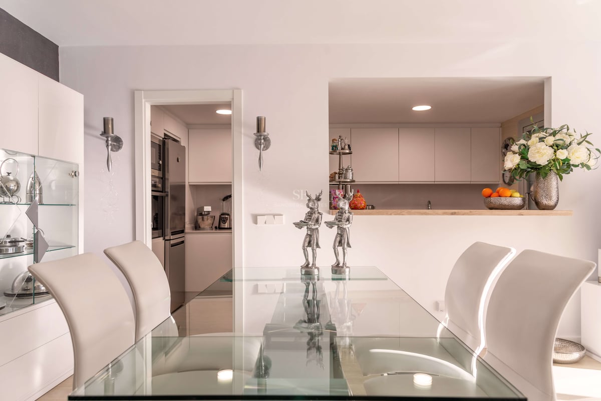 Zeitgenössisches Esszimmer mit Glastisch, weißen Stühlen und Blick in eine moderne weiße Küche durch eine Durchreiche.