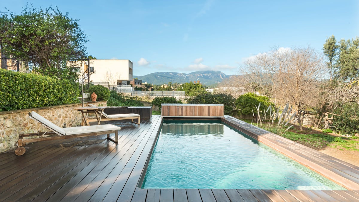 Rektangulär pool med mörkt trädäck, vita solstolar och panoramautsikt över bergen.