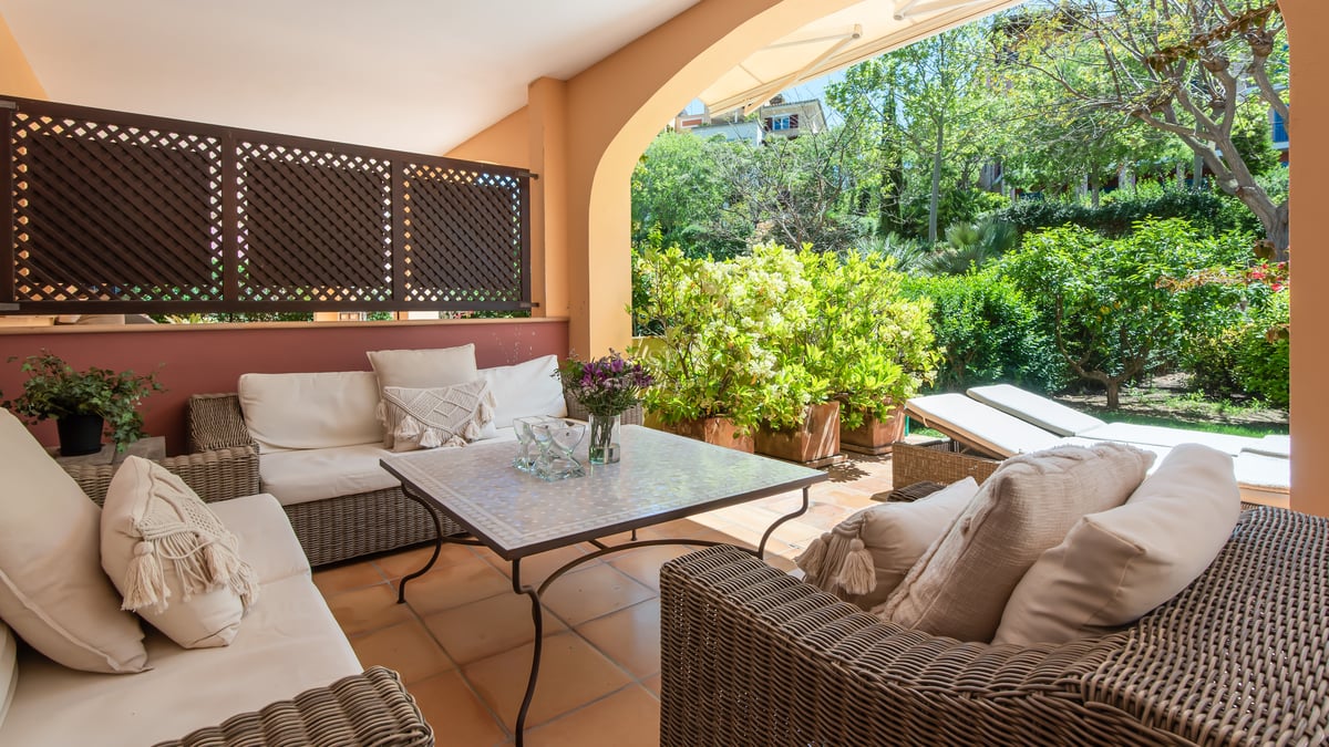 A covered terrace with wicker sofas, cream cushions, and a mosaic table overlooking a vibrant green garden.