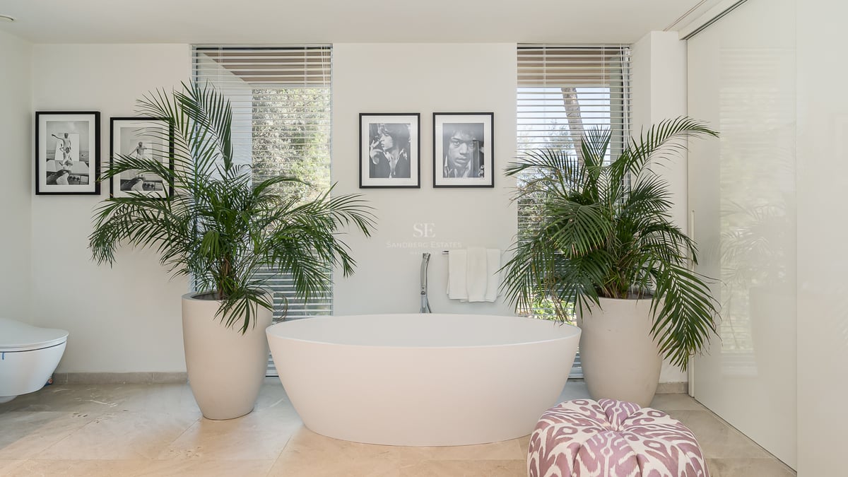 Freistehende weiße Badewanne flankiert von zwei großen Palmen vor bodentiefen Fenstern mit Jalousien.
