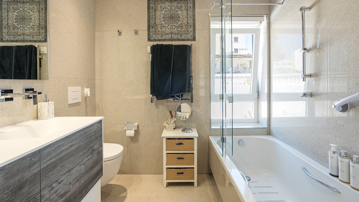 Modernt badrum med vitt badkar, glasskärm, kommod i träimitation och neutralt kakel med stenskänsla.