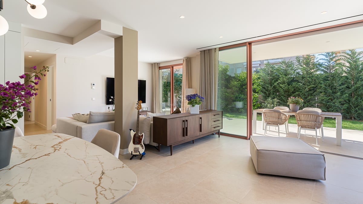 Modern living room with marble dining table, wood sideboard, and floor-to-ceiling glass doors opening to a garden.