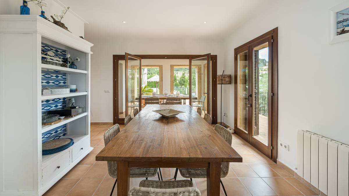 Intérieur avec grande table en bois, chaises en osier, sol en terre cuite et portes vitrées ouvrant sur une terrasse.