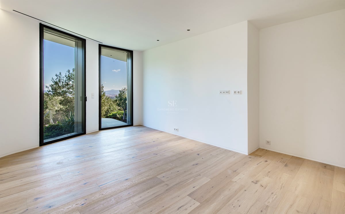 Une pièce vide avec du parquet en bois clair, des murs blancs et deux grandes baies vitrées donnant sur des arbres verts.