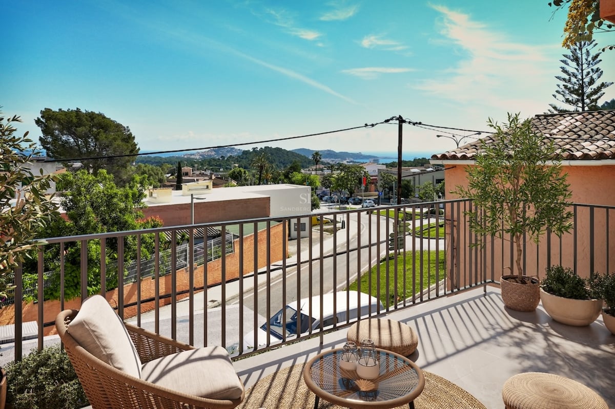 Sunlit terrace with wicker furniture overlooking a coastal town and the sea under a clear blue sky.
