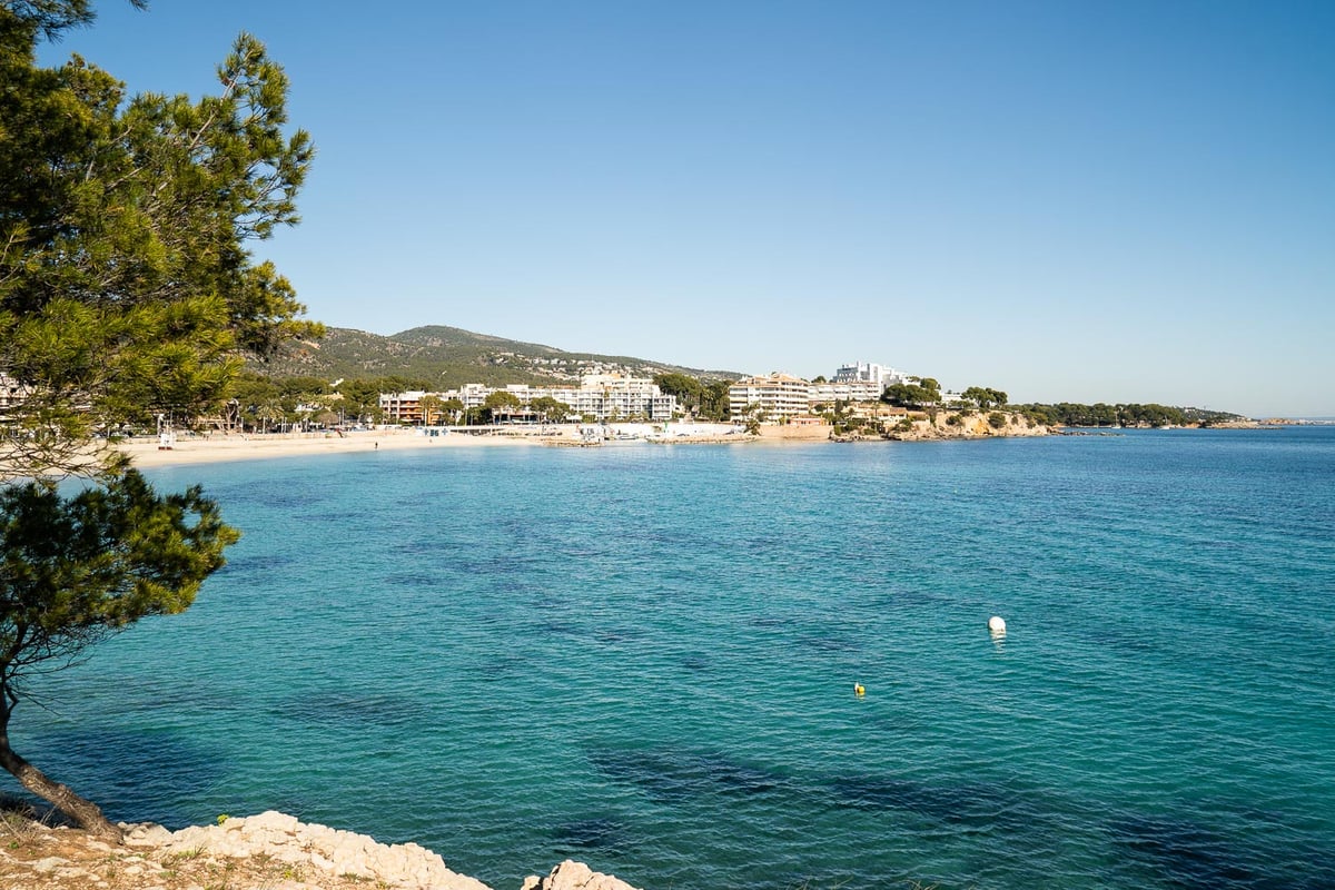 Weitläufiger Blick auf eine mediterrane Bucht mit klarem türkisfarbenem Wasser, Sandstrand und weißen Gebäuden.