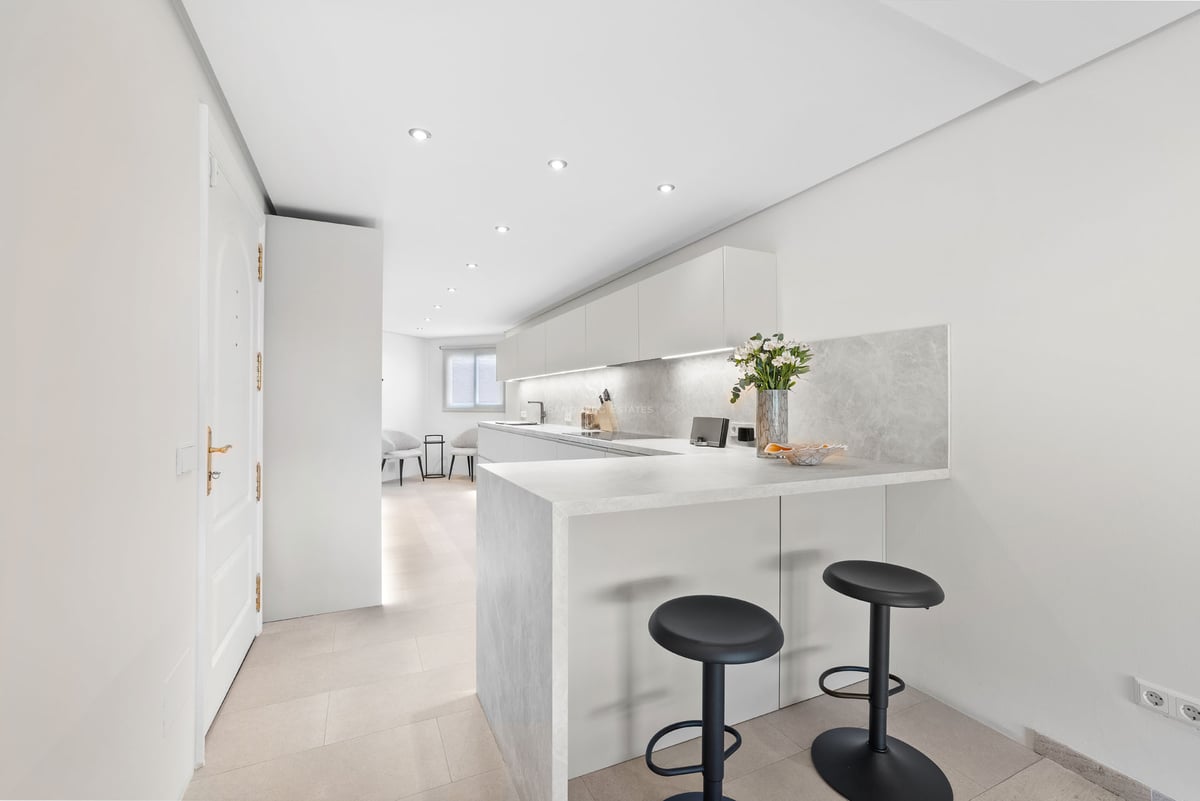 Une cuisine blanche moderne avec un comptoir en pierre, deux tabourets noirs et un éclairage LED intégré.